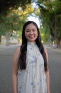 A young asian woman with long dark brown hair wears a high-collared sleeveless white dress with small blue floral details and smiles at the camera. 